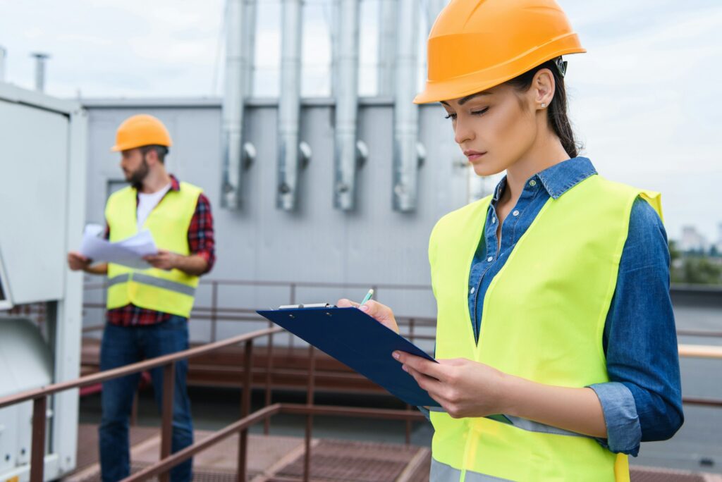 female architect in helmet writing in clipboard on roof, male colleague with blueprint behind